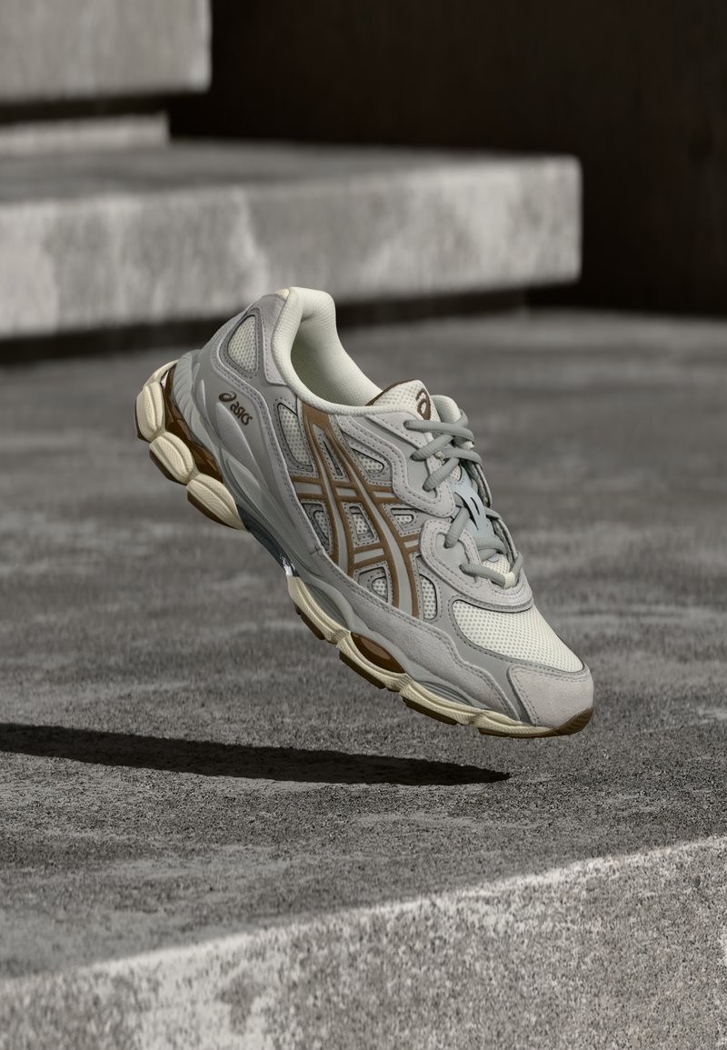 Ανοιχτό γκρι αθλητικό παπούτσι για τρέξιμο με δίχτυ από πάνω και καφέ λεπτομέρειες, διαθέτοντας μαξιλαρωτή σόλα και υφασμάτινη εξωτερική σόλα από καουτσούκ.