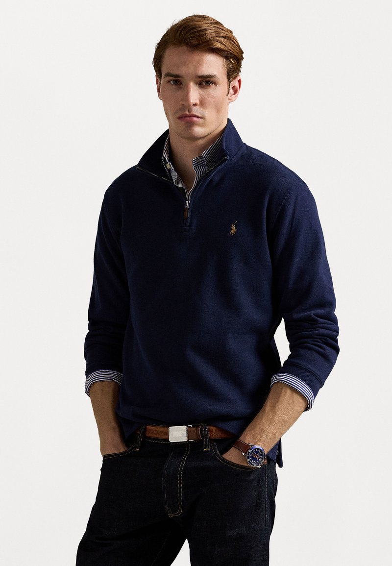 Young man with hands in pockets wearing a navy quarter-zip sweater over a striped shirt and dark jeans with a brown belt and watch.