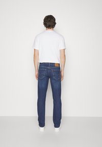 Tmavě modré džínové kalhoty s volnějším střihem, vybavené zadními kapsami a světle hnědou koženou značkou na pase. Nošeny s bílým tričkem.