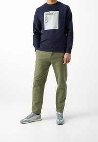 Marinblå sweatshirt med en ljusblå grafisk design och text, kombinerad med olivgröna avsmalnande byxor och grå träningsskor.