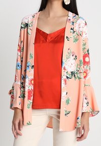 Blommig kimonojacka i persika med långa ärmar. Har livfulla blommor i rött, blått och grönt. Matchas med en röd linne.