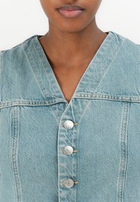 Lyseblå denimskjorte med dyp V-hals, prydet med fire sølvknapper og synlige sømdetaljer, som gir en teksturert finish.