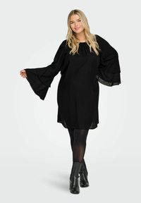 Crna haljina s zvonastim rukavima, teksturiranim materijalom i slobodnim krojem, uparena s crnim gležnjačama i tajicama. Jednostavan, elegantan dizajn.