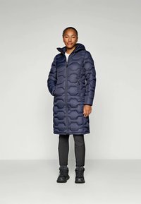 Marineblå pufferjakke med en teksturert, bølgete mønster, glidelås foran og hette. Brukt med svarte leggings og mørke sko.