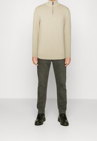 Béžový svetr na zip s dlouhými rukávy, v kombinaci s tmavě šedými kalhotami a černými šněrovacími botami. Jemná textura a přiléhavý střih.