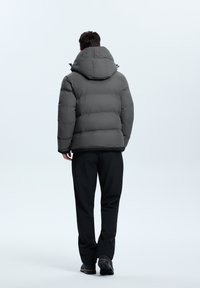 Graue gepolsterte Jacke mit großer Kapuze, geprägtem Logo auf dem Rücken und elastischen Bündchen; kombiniert mit schwarzen Hosen und robusten Schuhen.