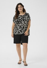 Top negro con flores blancas y rosas, de corte holgado, con mangas cortas. Combinado con pantalones cortos negros y sandalias negras de tira. Fondo plano.