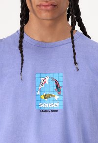 Camiseta de algodón morada con un gráfico de peces koi y texto "sensei" en blanco, sobre un fondo de rejilla. Mangas cortas y cuello redondo.