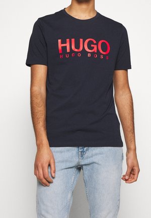 Navy blue cotton t-shirt featuring bold red "HUGO HUGO BOSS" text. Short sleeves, crew neck, slim fit. Light blue denim jeans visible.