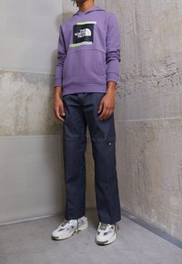 Sudadera con capucha morada y gráfico del logo en el frente, combinada con pantalones oscuros grises y zapatillas blancas con acentos verdes.