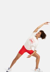 T-shirt en coton blanc avec motif rouge, associé à un short rouge en stretch. Le modèle est en position d'étirement sur le côté, portant des baskets blanches et bleues.