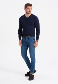 Marinblå V-hals tröja i ett mjukt material, kombinerad med slim fit blå jeans och mörkbruna mockaloafers. Minimalistisk klocka på vänster handled.