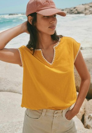 Young woman in yellow sleeveless top and pink cap stands by rocky shore with ocean waves in the background, hand resting on neck.