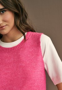 Fuchsia ull ärmlös tröja med ribbad halsringning lager på lager över en vit kortärmad skjorta. Mjuk textur, avslappnad passform.
