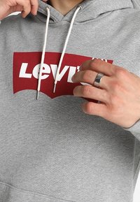 Sudadera gris con un logo rojo de Levi's. Cuenta con una capucha con cordones blancos y una textura suave. Manos visibles ajustando la prenda.