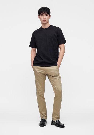 Jeune homme debout, les mains dans les poches, portant un t-shirt noir, un pantalon beige et des chaussures habillées noires sur un fond blanc uni.