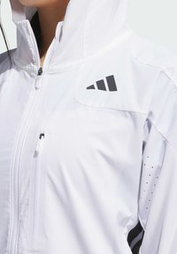 Veste zippée blanche fabriquée dans un matériau léger et texturé. Elle présente un col haut, une poche poitrine zippée et un logo noir sur l'épaule.