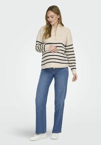 Beige gestrickter Pullover mit navyfarbenen horizontalen Streifen, halblangem Reißverschluss am Hals, langen Ärmeln, kombiniert mit blauen geraden Jeans und weißen Turnschuhen.