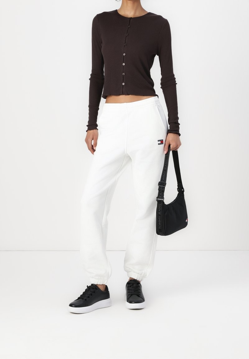 Femme portant un top court noir à manches longues boutonné, pantalon de jogging blanc avec logo, baskets noires, tenant un petit sac à bandoulière noir sur fond blanc.