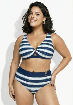 Donna sorridente con capelli castani medi, che indossa un costume da bagno a due pezzi bianco e blu a righe con dettagli ad anello, posando su sfondo bianco.