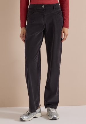 Personne portant un pantalon noir droit, un haut rouge à manches longues et des baskets argentées, debout devant un fond uni.