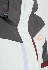 Kurtka wiatrówka złożona z paneli w odcieniach szarości, jasnej mięty i bordo. Gładka zewnętrzna faktura z kieszenią na zamek i detalem z logo.