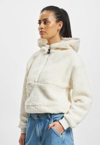 Hvit fleece-hettejakke med glidelås foran, elastiske mansjetter og stor hette, brukt sammen med høyhalsa blå jeans. Myk, teksturert stoff.