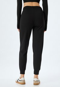 Sorte tapered joggers med elastisk talje og manchetter ved anklerne, lavet af blødt stof, parret med hvide sneakers med sorte detaljer.
