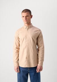 Chemise beige à manches longues avec boutonnières, en matériau coton, col pointu, logo brodé sur la poitrine, associée à un jean en denim bleu.