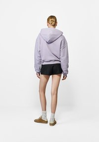 Lila oversized kapucnis pulóver fekete sorttal párosítva. A modellen bézs cipők és fehér zoknik vannak, hátulról nézve, fehér háttér előtt.