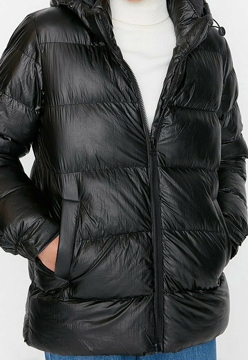 Personne portant une doudoune noire brillante avec capuche, fermeture éclair et poches latérales, par-dessus un pull à col roulé blanc.