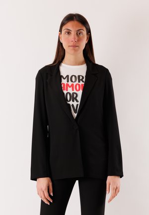Donna con capelli lunghi castani che indossa un blazer nero sopra una camicia bianca con la scritta "AMOR" in testo rosso e nero, in piedi contro uno sfondo chiaro e uniforme.