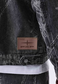 Chaqueta de mezclilla en negro con detalles desgastados. Presenta una etiqueta de cuero marrón con el logo "PXP" y herrajes plateados. Superpuesta sobre una camisa blanca.