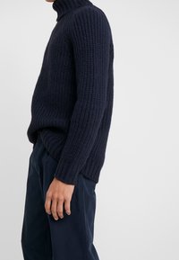 Homme portant un pull à col roulé côtelé bleu marine et un pantalon bleu foncé, debout devant un fond clair uni.