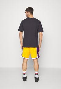 T-shirt nera a maniche corte abbinata a pantaloni da basket gialli con dettagli viola e rifiniture bianche, indossata con calze bianche e nere.