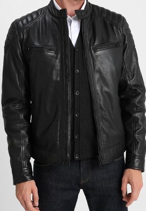 Chaqueta de cuero negra con acolchado acolchado en los hombros, bolsillos en el pecho con cremallera, llevada sobre un cárdigan oscuro abotonado y una camisa blanca con cuello.