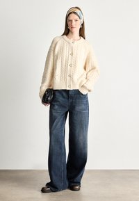 Cárdigan de punto color marfil con patrones de cable, con cuello redondo y botones perla, combinado con pantalones vaqueros de pierna ancha en denim oscuro. Accesorio de bolso de mano negro.