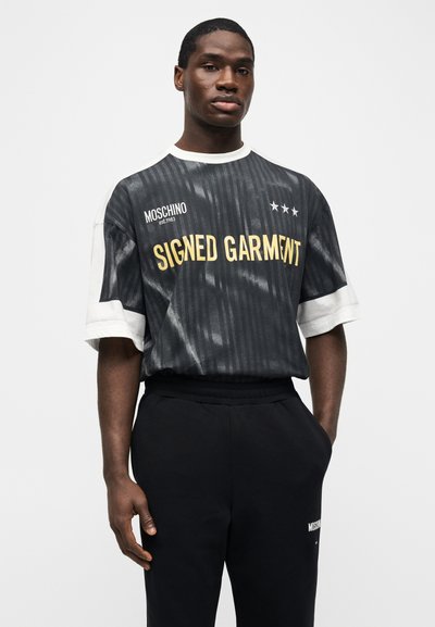 Άνδρας που φοράει μαύρο και άσπρο κοντομάνικο πουκάμισο με την επιγραφή "SIGNED GARMENT" και μαύρο παντελόνι με τα χέρια στις τσέπες, στέκεται μπροστά σε απλό λευκό φόντο.