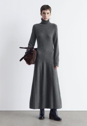 Frau mit kurzen Haaren, die ein langes graues Rollkragenkleid, schwarze Stiefel, Ringohrringe trägt und eine große braune Handtasche hält.