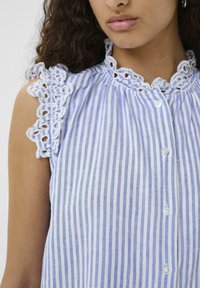 Blusa a righe blu e bianche con dettagli in pizzo sulle spalle, caratterizzata da un collo rotondo e chiusure a bottoni lungo la parte anteriore.