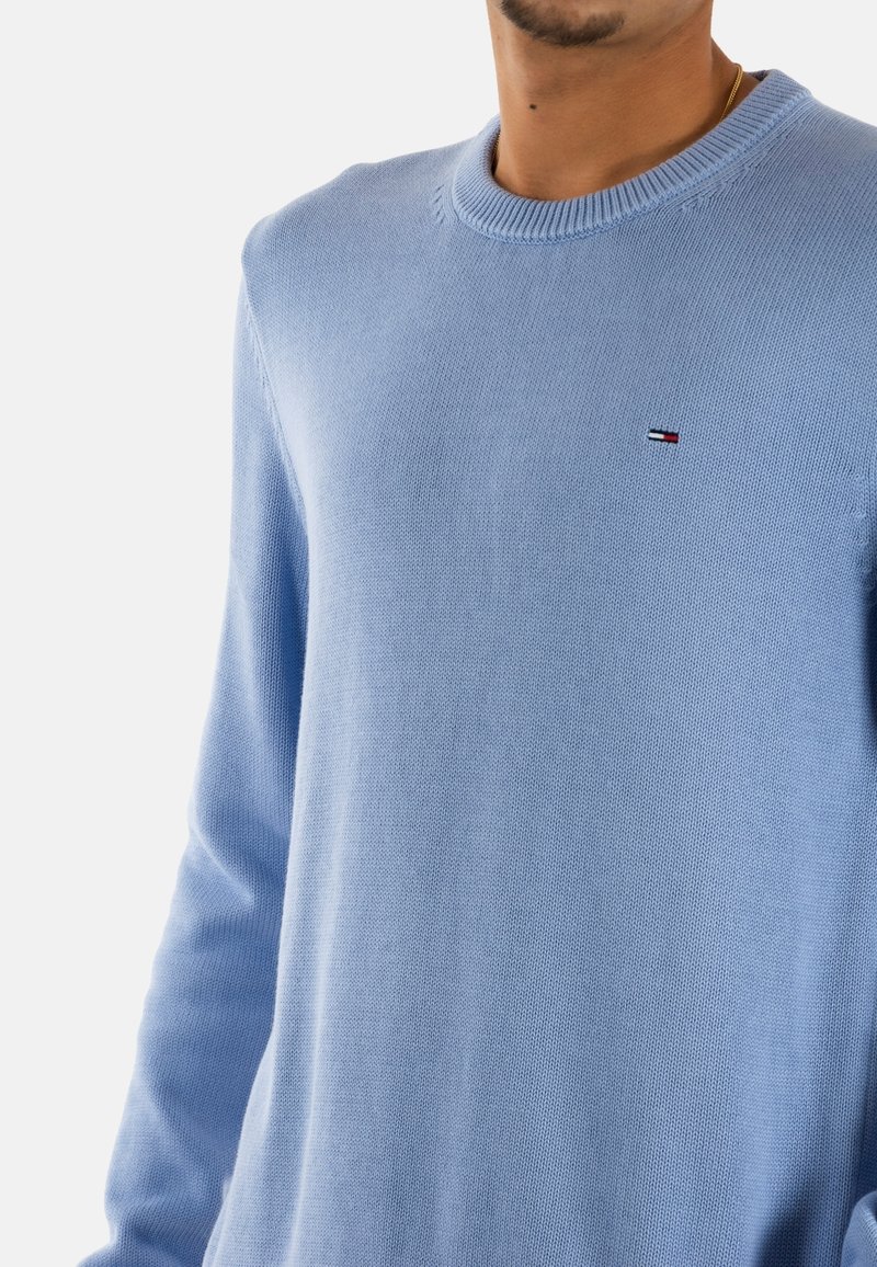 Lichtblauwe sweater van zacht breiwerk, met een klassieke ronde hals en een klein logo in rood, wit en blauw.
