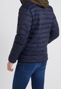 Navy puffer jacket with horizontal stitching, padded texture, and a brown hood. Features a logo detail on the left sleeve. Partial view from the back.