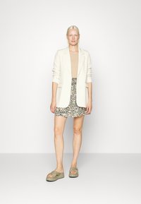 Blazer creme, blusa bege, saia mini floral em cores escuras, e sandálias knotted em verde-oliva. O blazer tem um corte descontraído e colarinho de lapela.