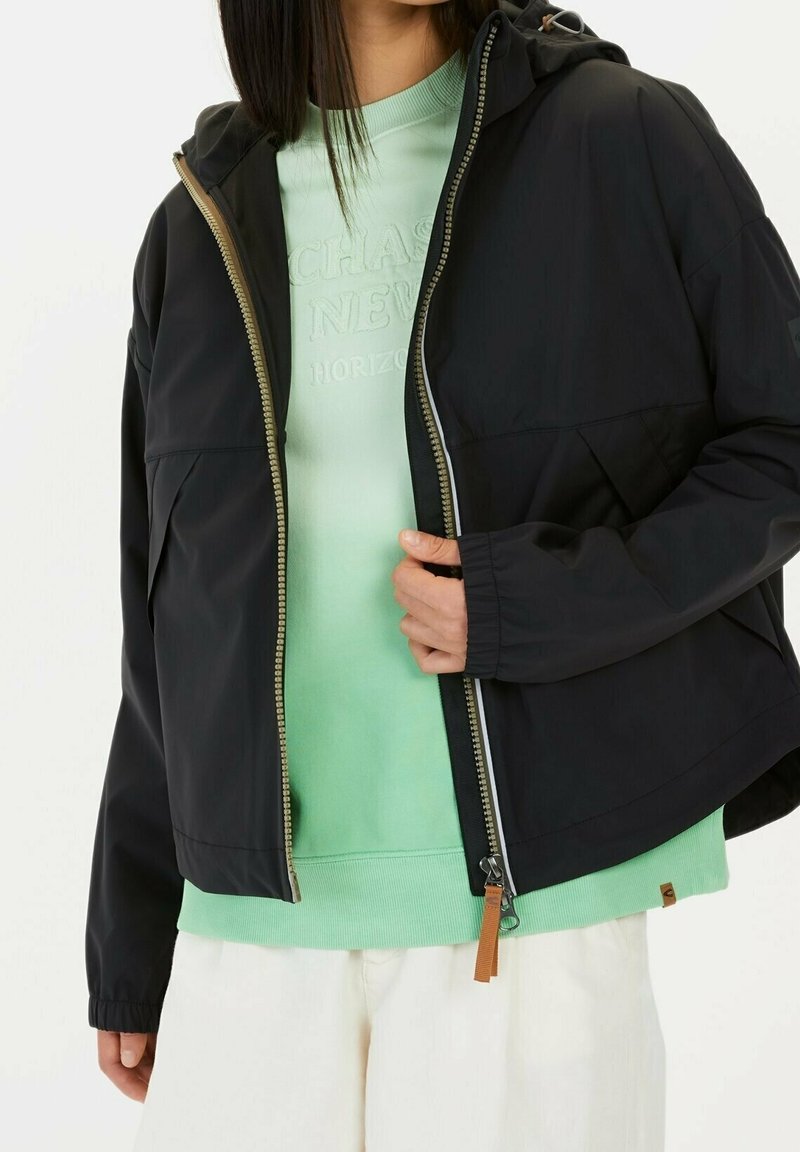 Personne portant une veste à capuche noire dézippée sur un sweat-shirt vert clair et un pantalon blanc, tenant la fermeture éclair de la veste d'une main.