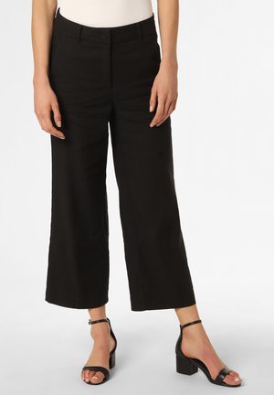 Frau trägt schwarze, weit geschnittene, knöchellange Hose, schwarze Sandalen mit Blockabsatz und Knöchelriemen sowie ein weißes, ärmelloses Oberteil, steht vor weißem Hintergrund.