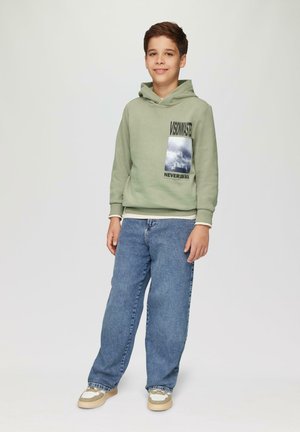 Bluza z kapturem w jasnym zielonym kolorze z graficznym nadrukiem, zestawiona z luźnymi niebieskimi dżinsami i beżowymi trampkami z białymi akcentami.