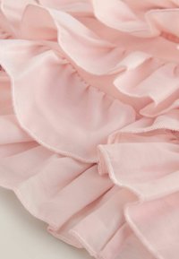 Hele roosa volangidega kangas pehme tekstuuriga; iseloomulikud pehmed lainelised servad ja nähtavad õmblused, mis loovad kihilise disaini.