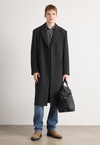 Grauer Wollmantel mit großen Revers, fünf Knöpfen und seitlichen Taschen, getragen über einem grauen Hemd, kombiniert mit blauen Jeans und braunen Schuhen.