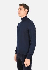 Maglione a collo alto blu navy realizzato in morbido tessuto a maglia. Design aderente con dettagli a coste sul colletto e sui polsini, abbinato a pantaloni scuri.