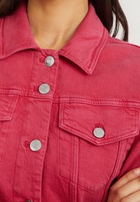 Červená džínsová bunda s límcem, stříbrnými druky a dvěma náprsními kapsami. Má mírně vybledlou texturu a viditelné detaily šití.